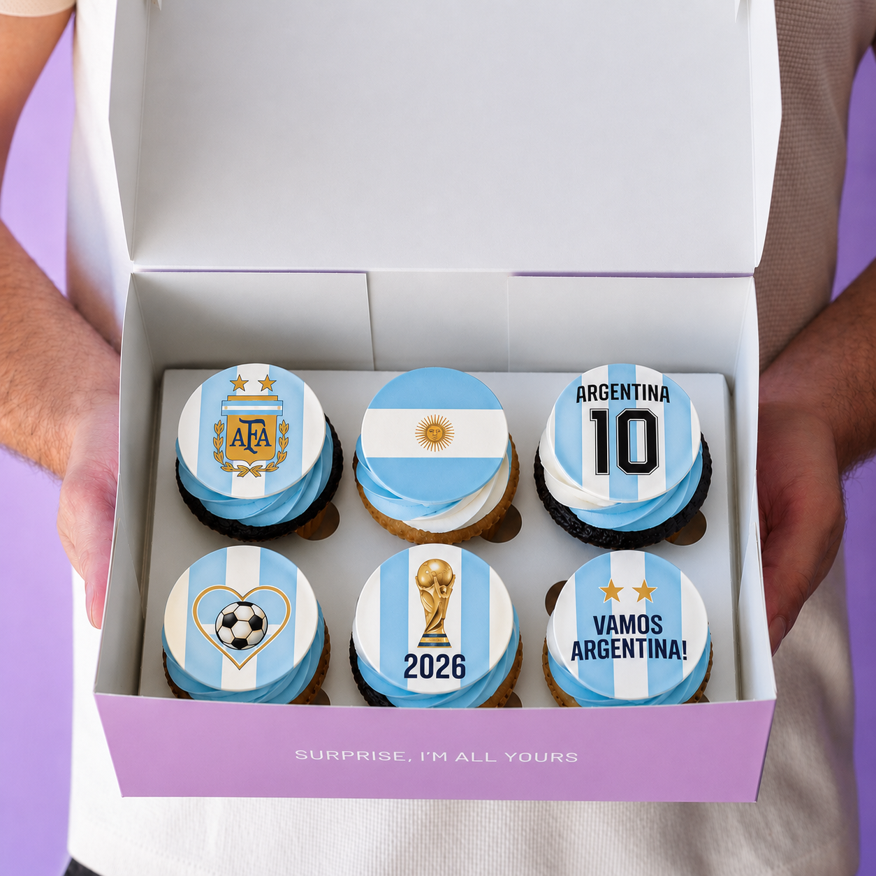 A box of six Argentina-themed cupcakes with light blue and white striped frosting, decorated with national team and soccer motifs, held open by two hands against a purple background.