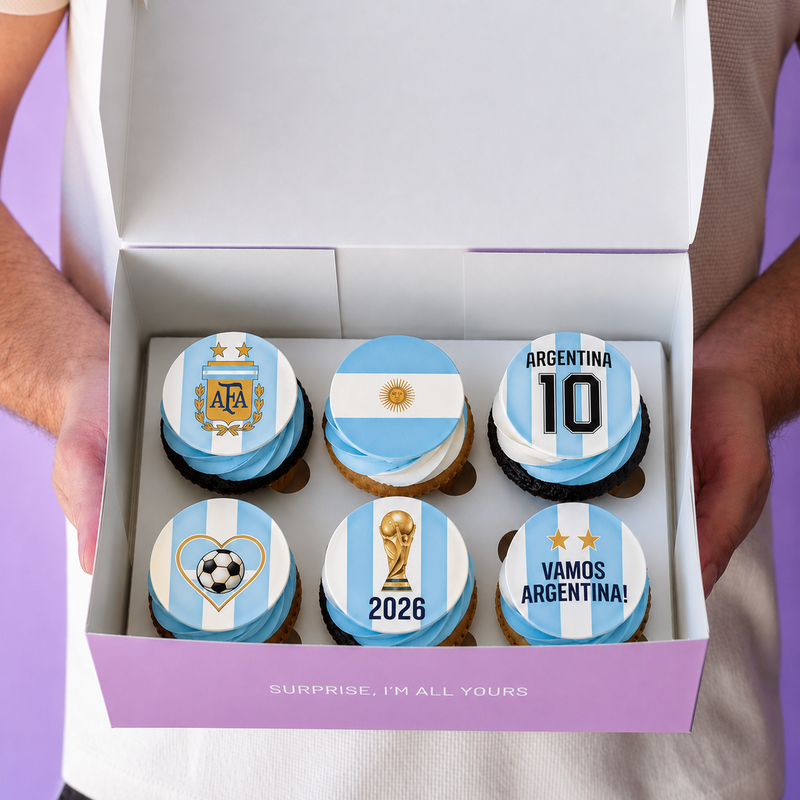 A box of six Argentina-themed cupcakes with light blue and white striped frosting, decorated with national team and soccer motifs, held open by two hands against a purple background.