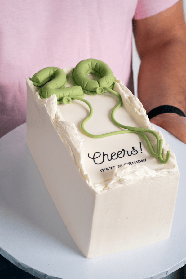 Small birthday cake with green decorations and 'Cheers!' text on a white plate.