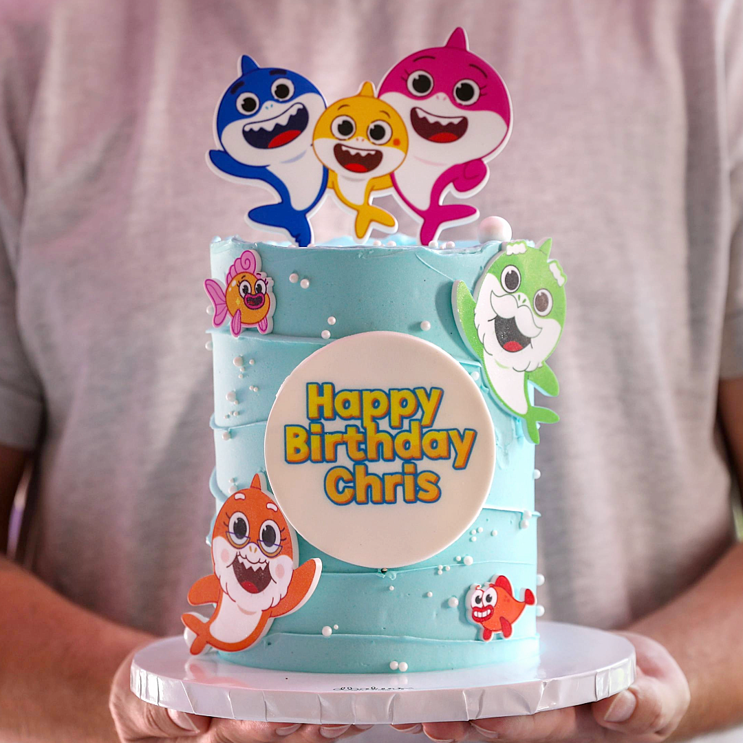 A colorful birthday cake decorated with characters from the Baby Shark song, with a 'Happy Birthday Chris' message on the front.