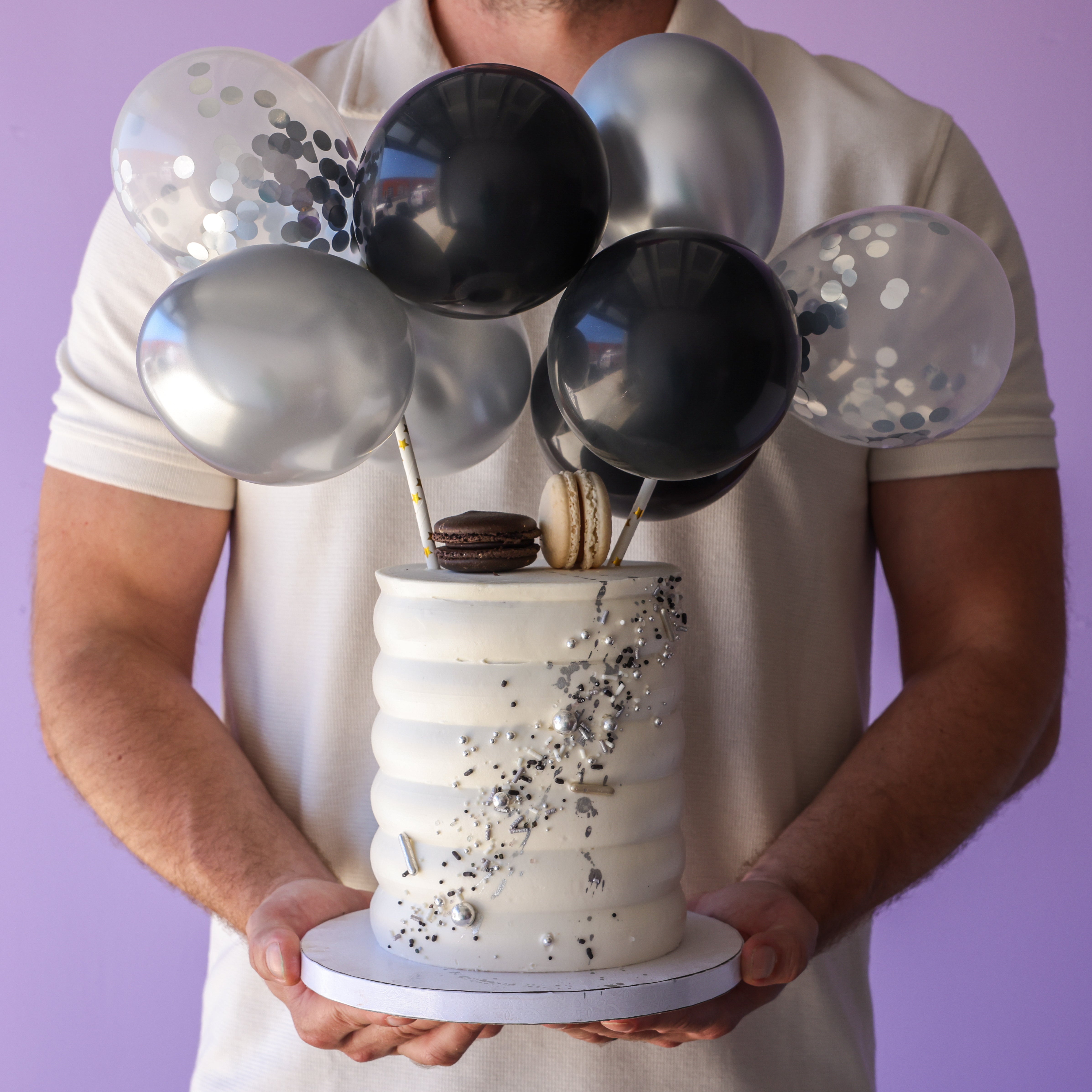 A person holding a birthday cake decorated with black and silver balloons and a wood dowel for structural support.