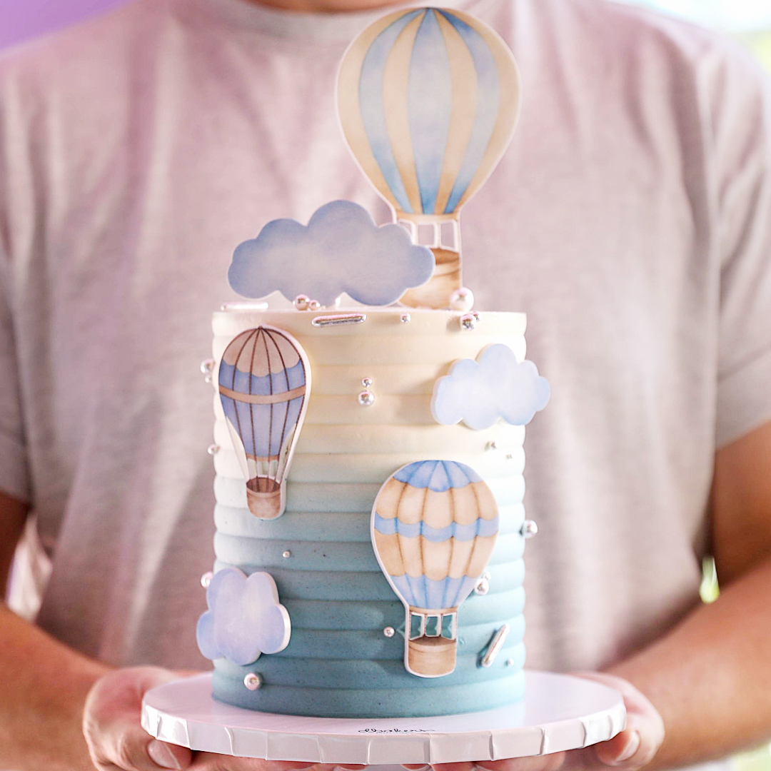A multi-layered cake decorated with hot air balloon and cloud figures in blue and white, held by a person.