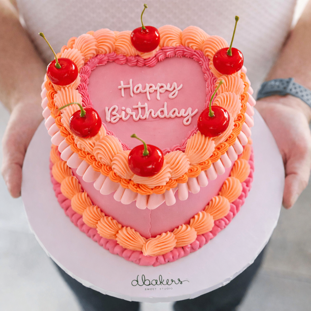 Heart-shaped birthday cake with pink and orange frosting, cherries, and 'Happy Birthday' text.