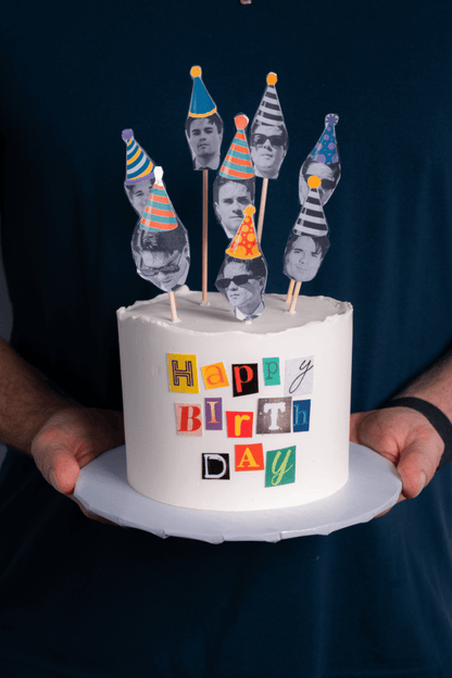 White birthday cake with colorful toppers and 'Happy Birthday' text, held by a person against a dark background.