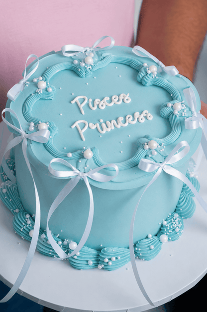 Light blue cake with 'Princess' text and decorative ribbons on a white plate.