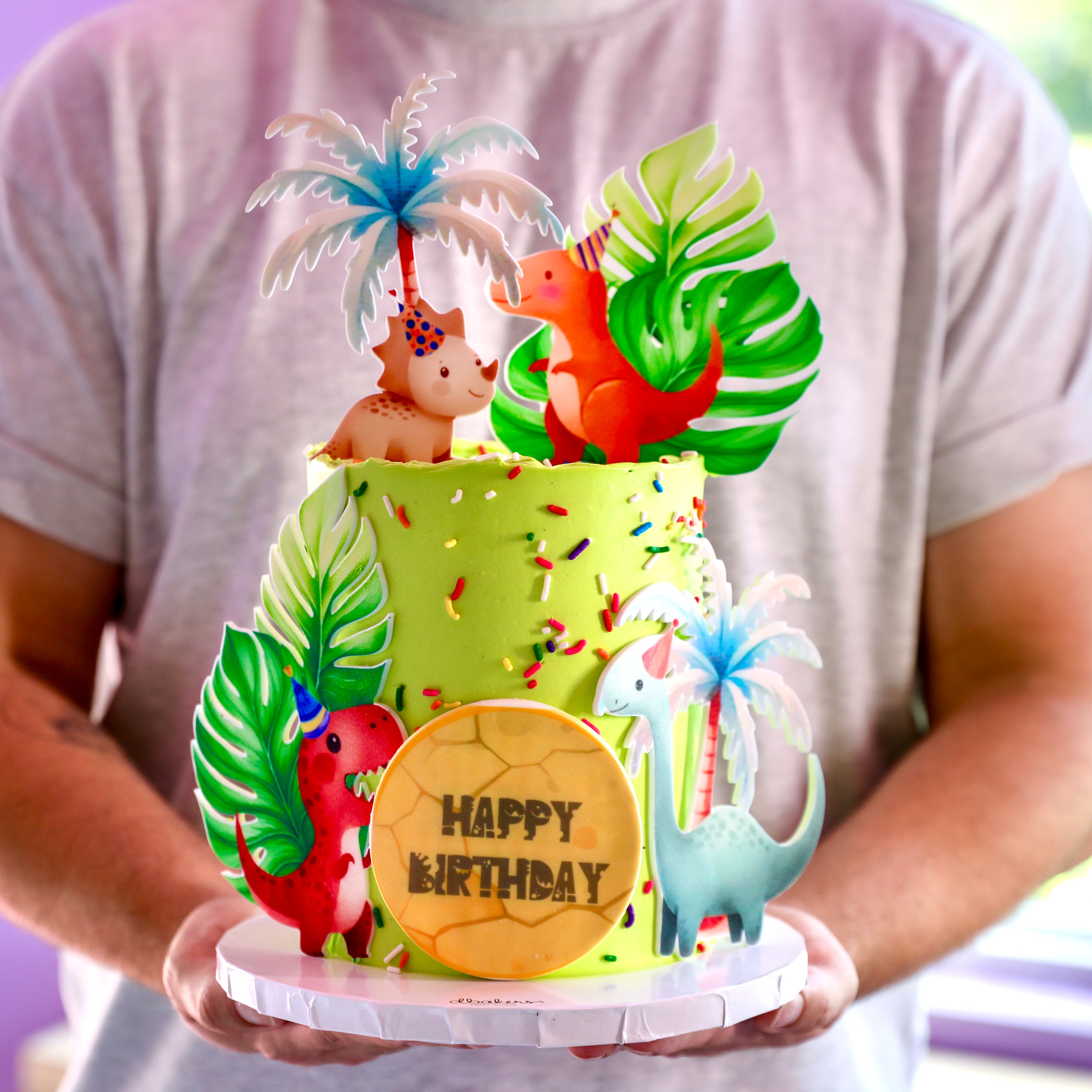 A colorful birthday cake decorated with dinosaur figurines, palm leaves, and a 'Happy Birthday' message on a plaque.