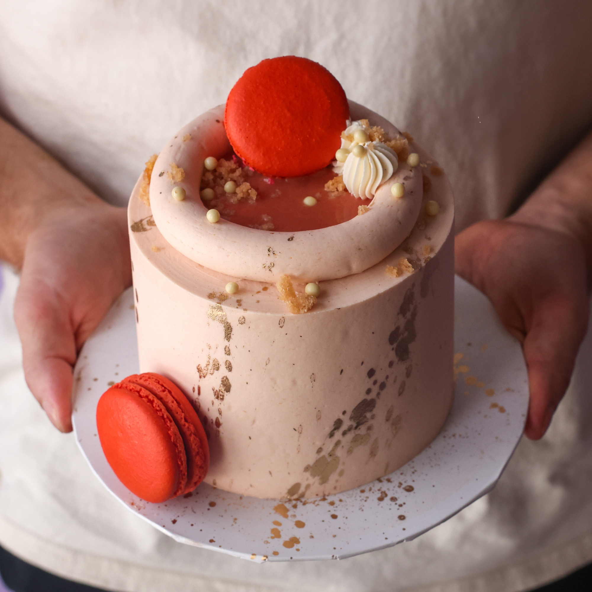 Small cake with red macarons and white cream on a white plate held by a person.