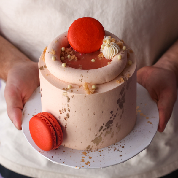 Small cake with red macarons and white cream on a white plate held by a person.
