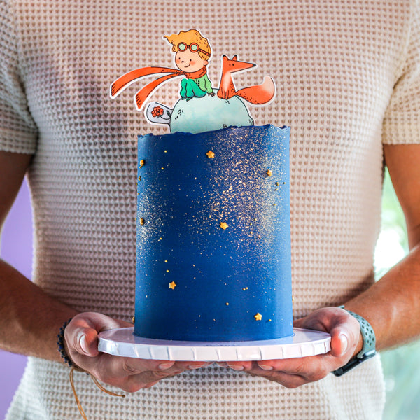 A person holding a small, blue round cake decorated with character figures from 'The Little Prince' story, with a sprinkling of gold stars on the icing.