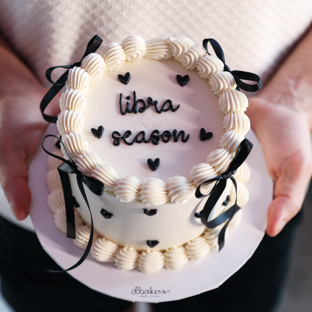 A round cake with white frosting and black decorations, with the words 'libra season' written on it, held in someone's hands.