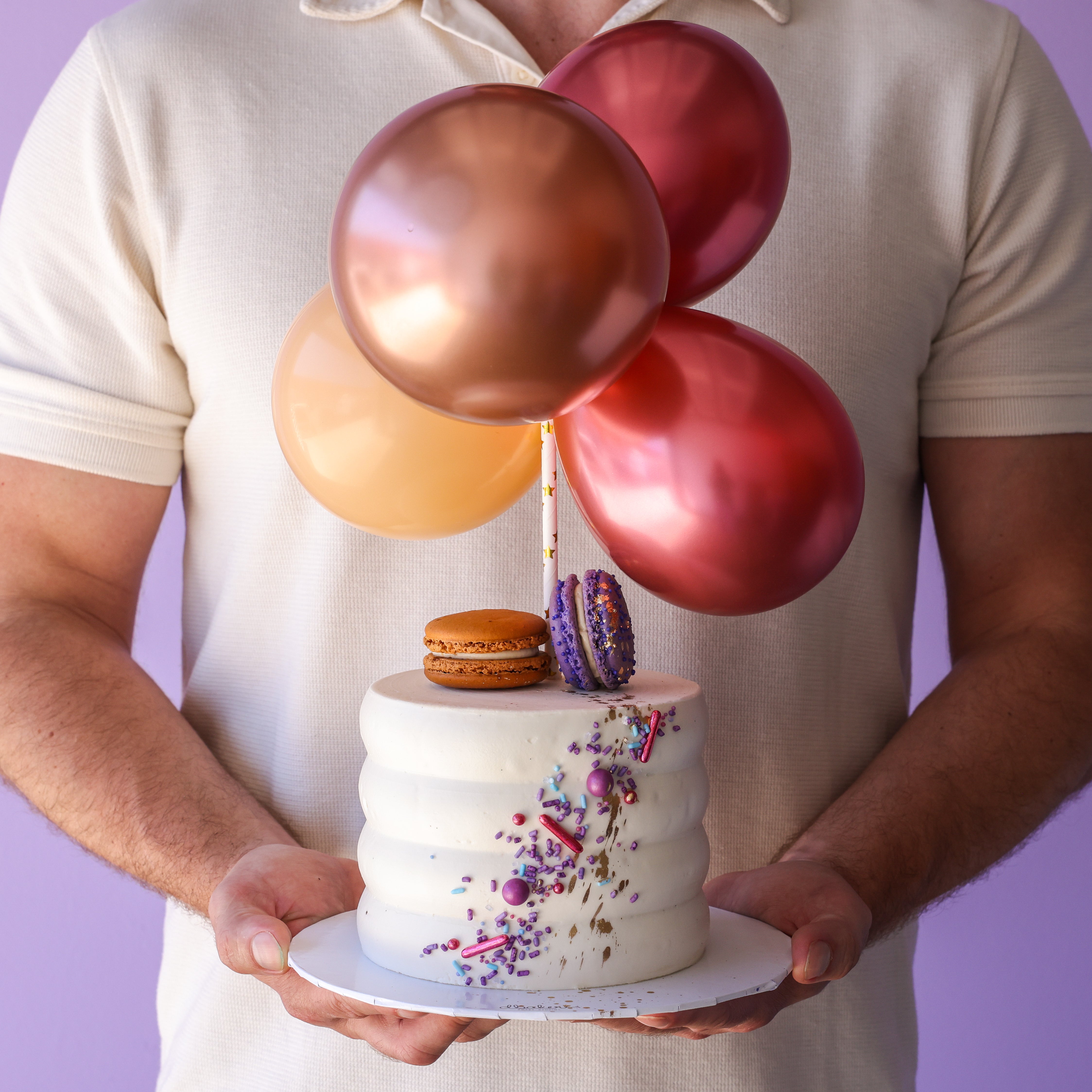 A person holding a birthday cake decorated with metallic pink and rose gold balloons and topped with a macaron.