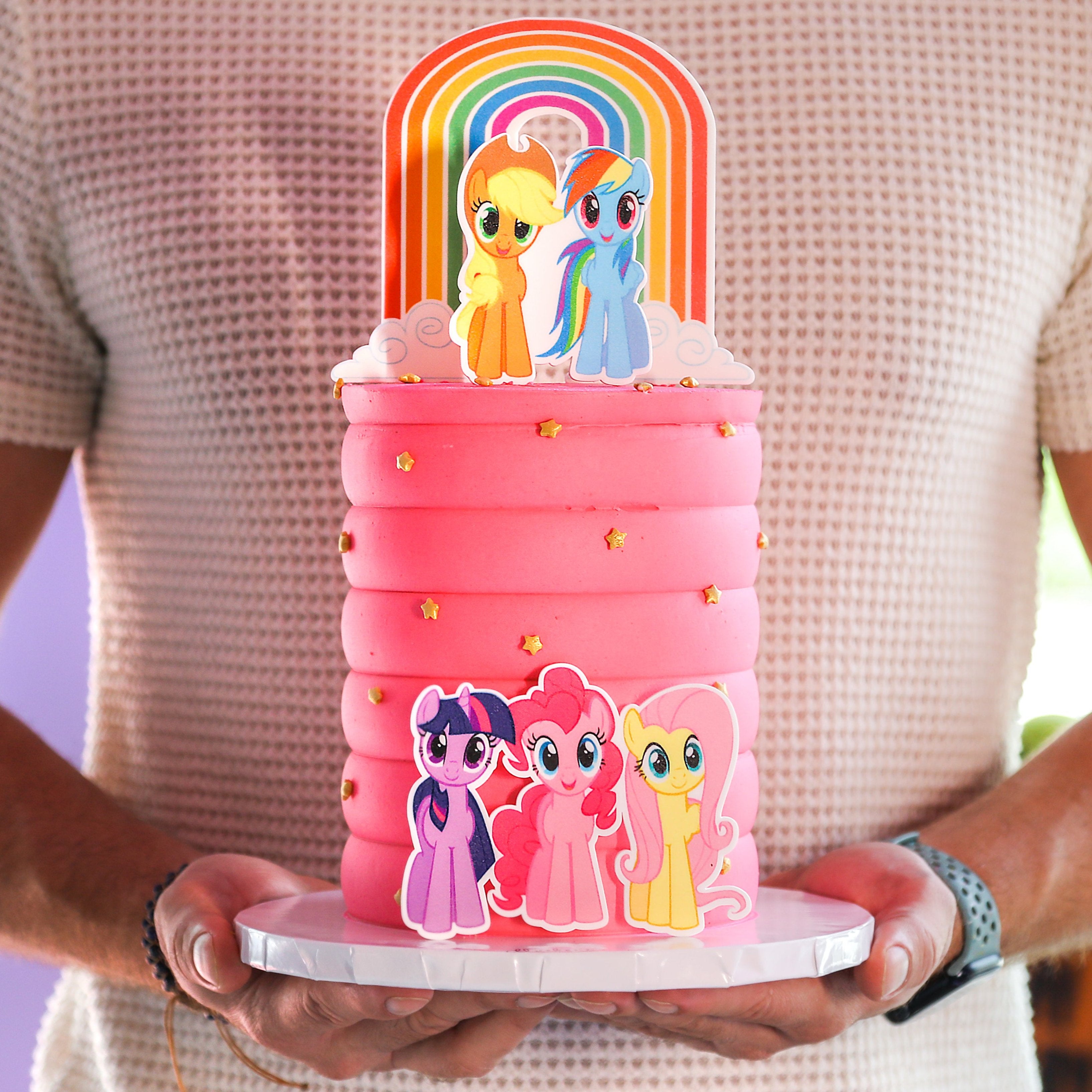 A cake decorated to look like the My Little Pony characters, with a pink base and multi-colored layers, held by a person.