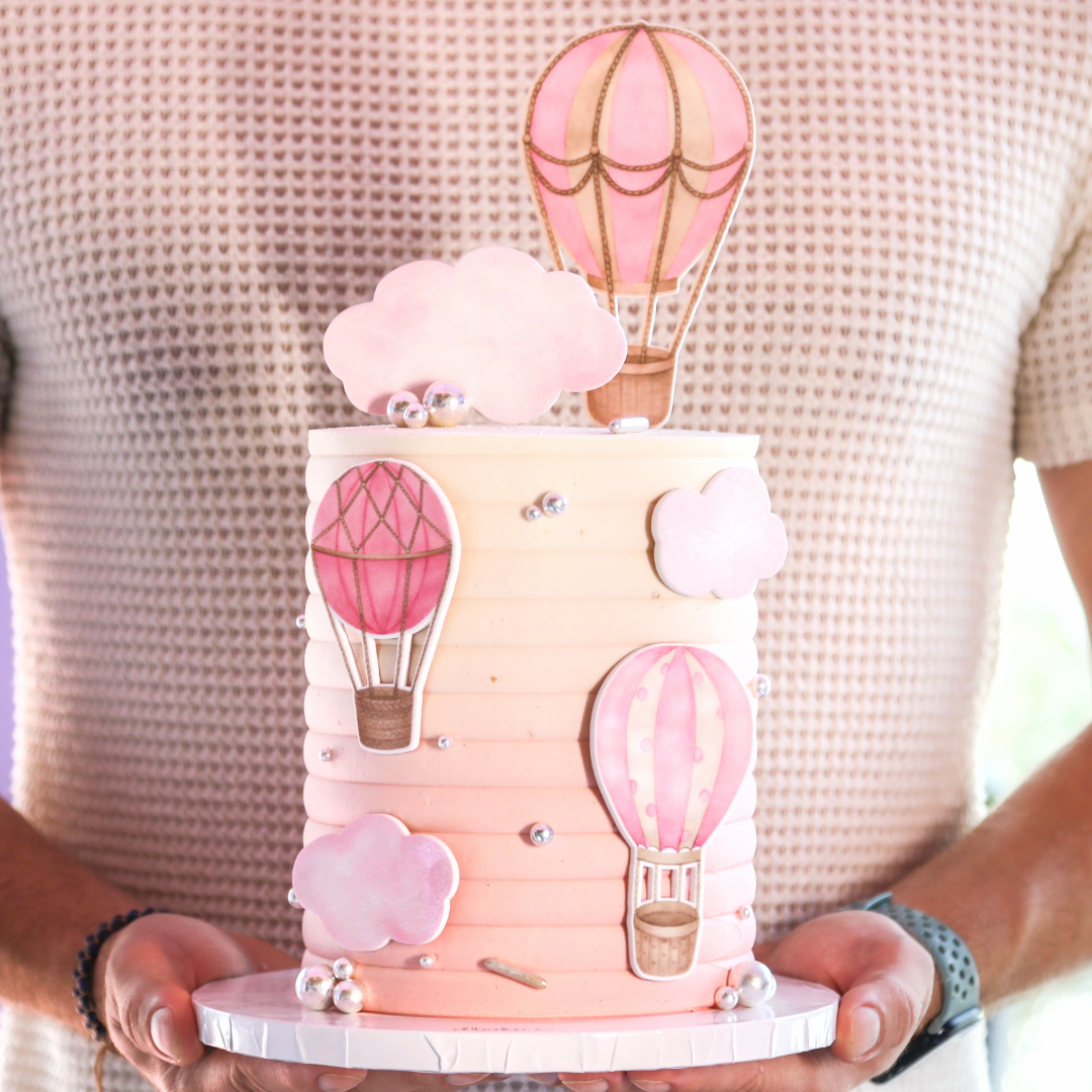 A pink-themed cake with balloon decorations and cloud accents, presented on a light background.