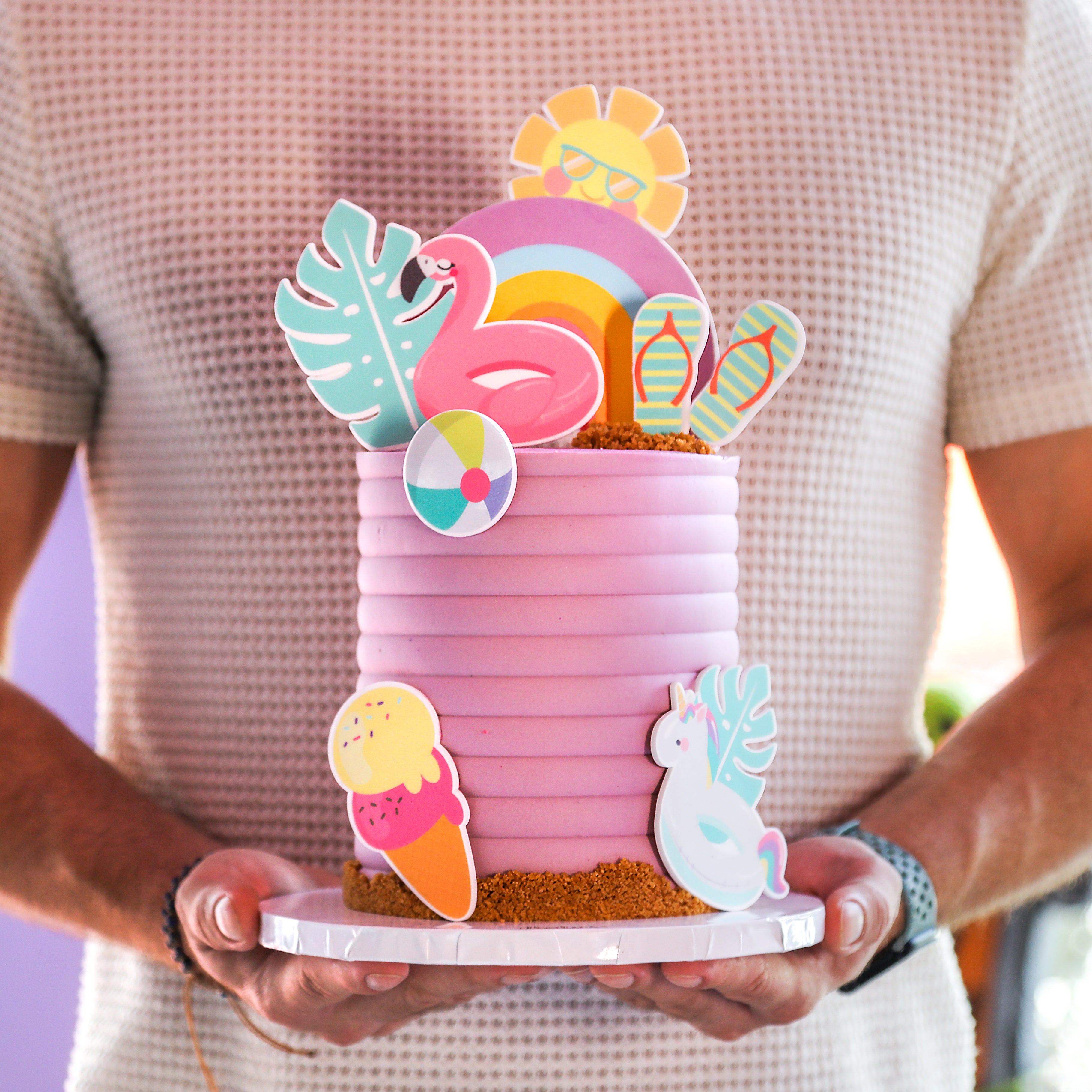 A person holding a decorated cake with pink layers and playful, colorful toppers including a flamingo, rainbow, and beach-themed items.