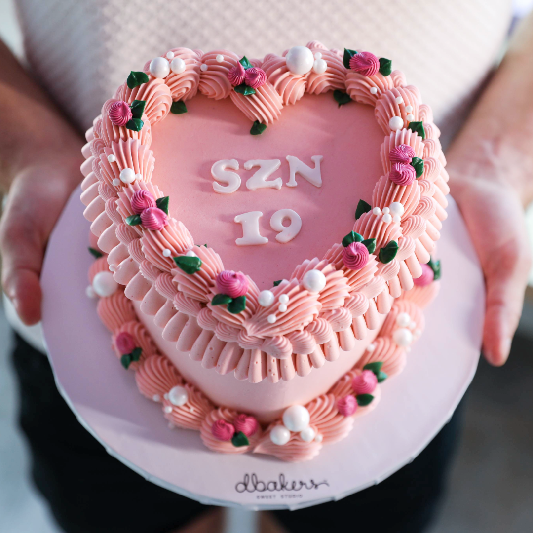 A heart-shaped cake decorated with pink rosettes and heart-shaped accents, with the text 'SZN 19' on top.