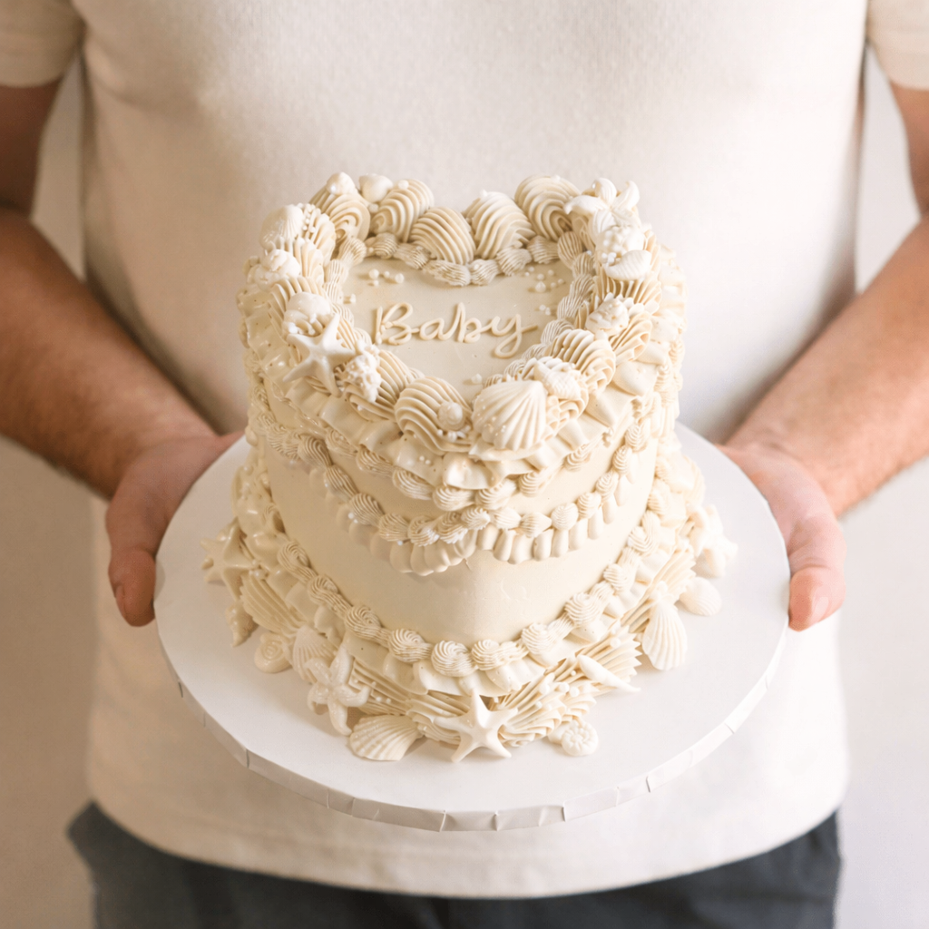 Person holding a decorative white cake with intricate designs and the word 'Baby' on a plain background