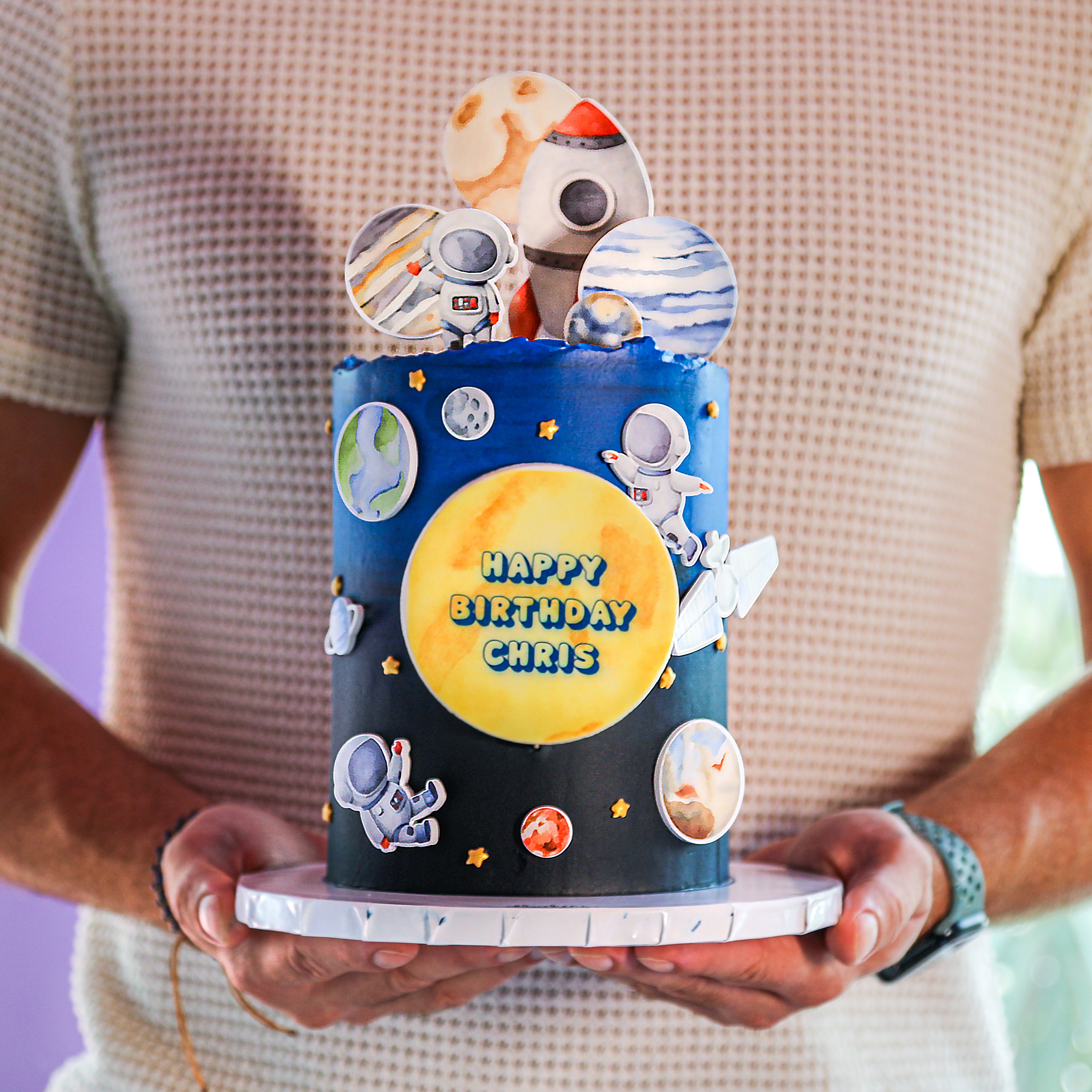 A decorated space-themed cake with astronaut figures, planets, and a 'Happy Birthday' message, held by a person.