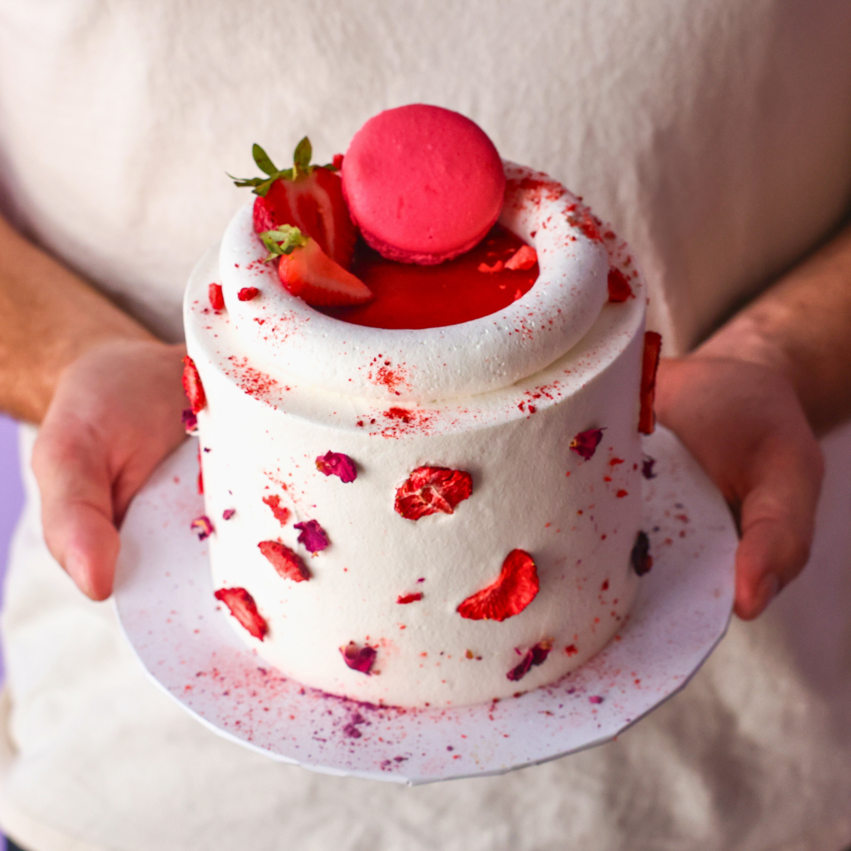 Person holding a small white cake with red decorations against a plain background
