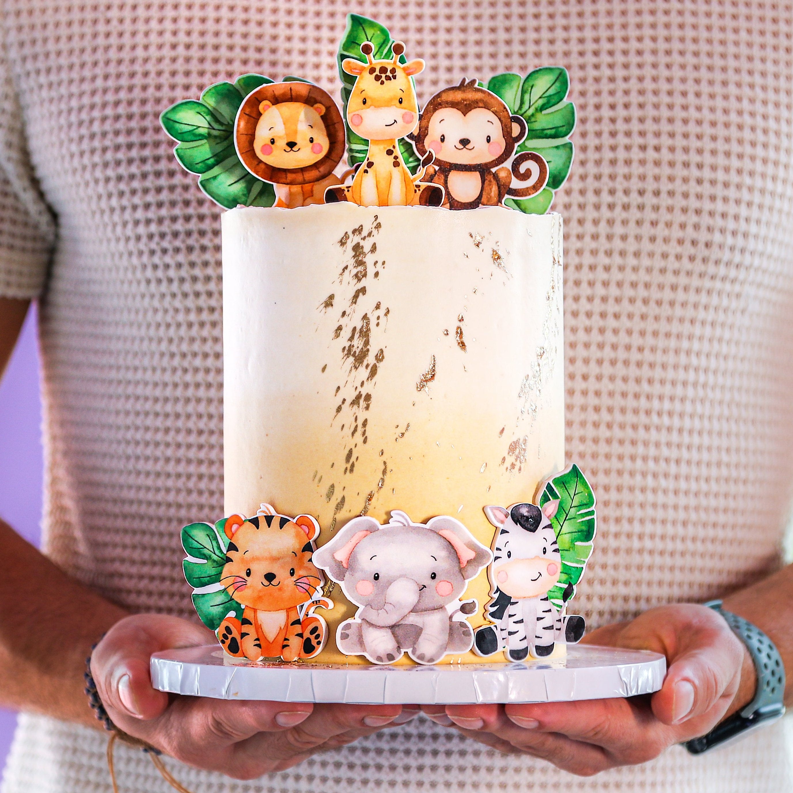 A decorative cake with animal figurines including a lion, monkey, and others, designed to look like a safari adventure theme. The cake is topped with green leaves and has a beige and white color scheme.